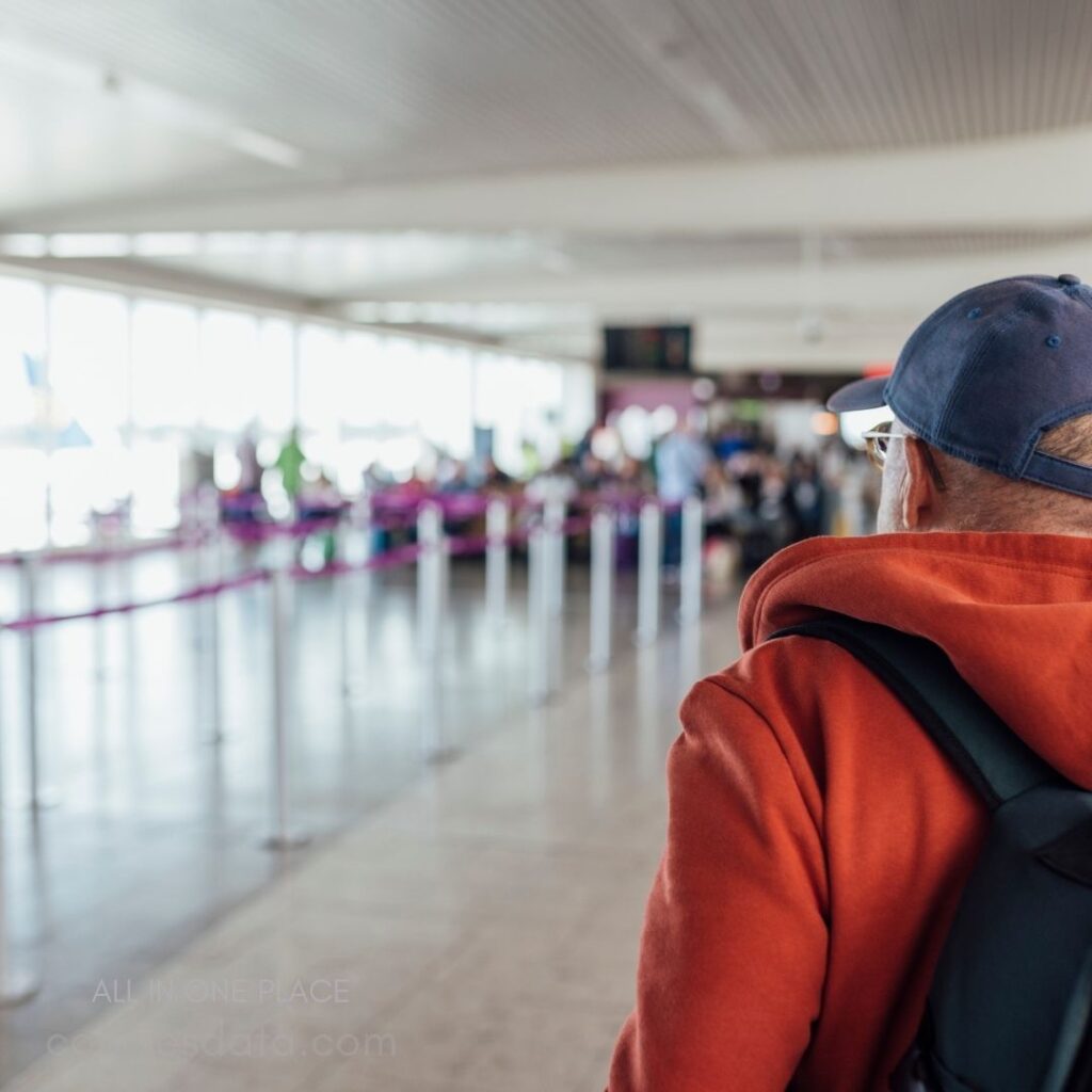 Man in orange hoodie waiting. Airport terminal background. Open space with queues. People in background. Soft natural lighting.