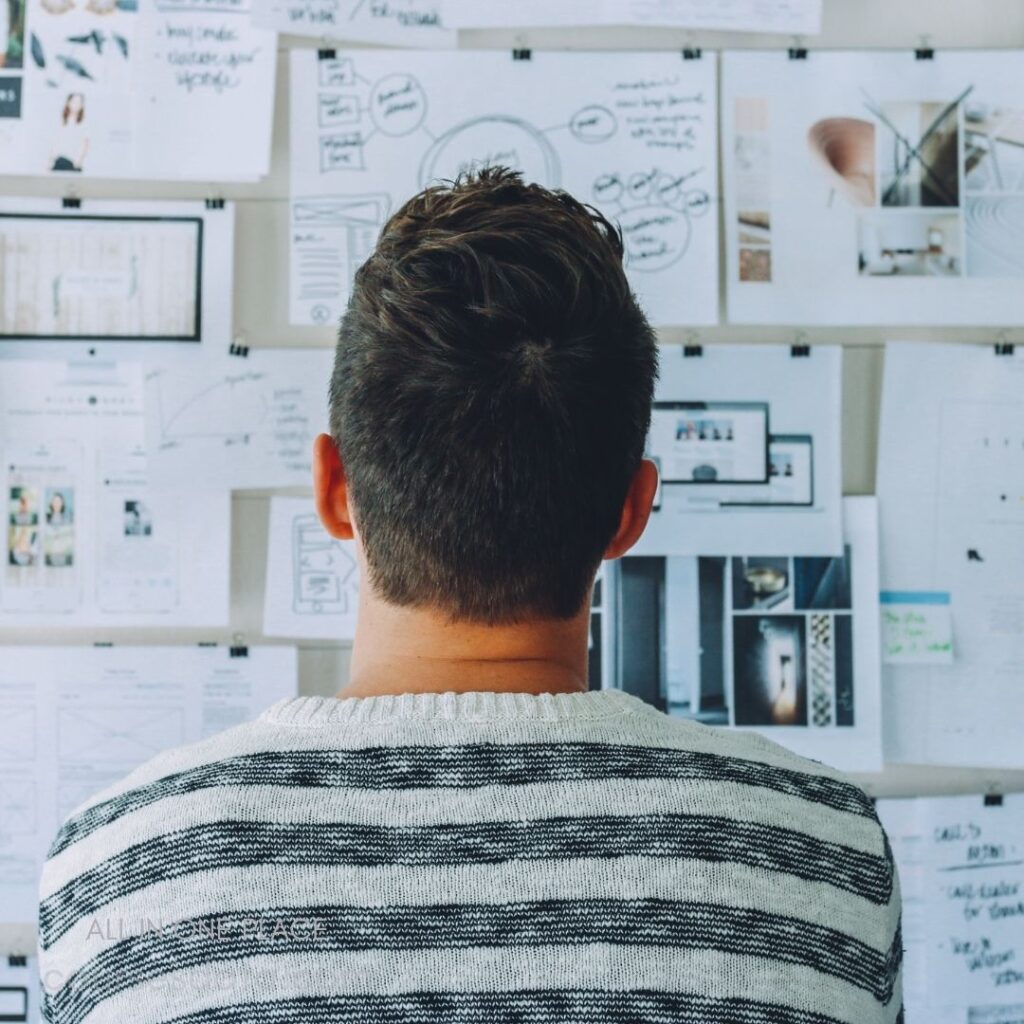 Person observing paper collage. Wall covered with design sketches. Stripped sweater, short hair visible. Notes and images pinned up. Creative brainstorming session depicted.