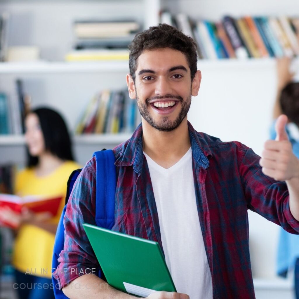 Smiling young man with books. Thumbs up gesture in library. Bookshelves filled with literature. Another person browsing behind him. Casual attire with backpack.