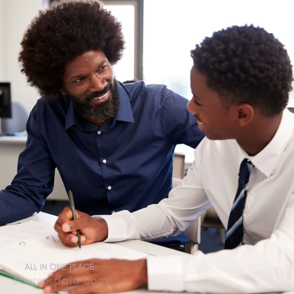 Smiling man mentoring young student. Discussion over notebook and pen. Bright, collaborative learning environment. Casual attire: blue shirt, tie.