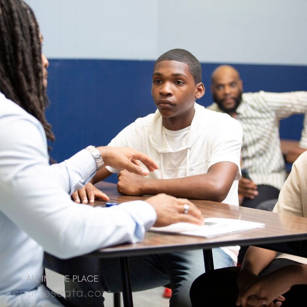 Two people in discussion. Young man listens intently. Table with papers in foreground. Background features another seated person. Classroom setting with blue walls.