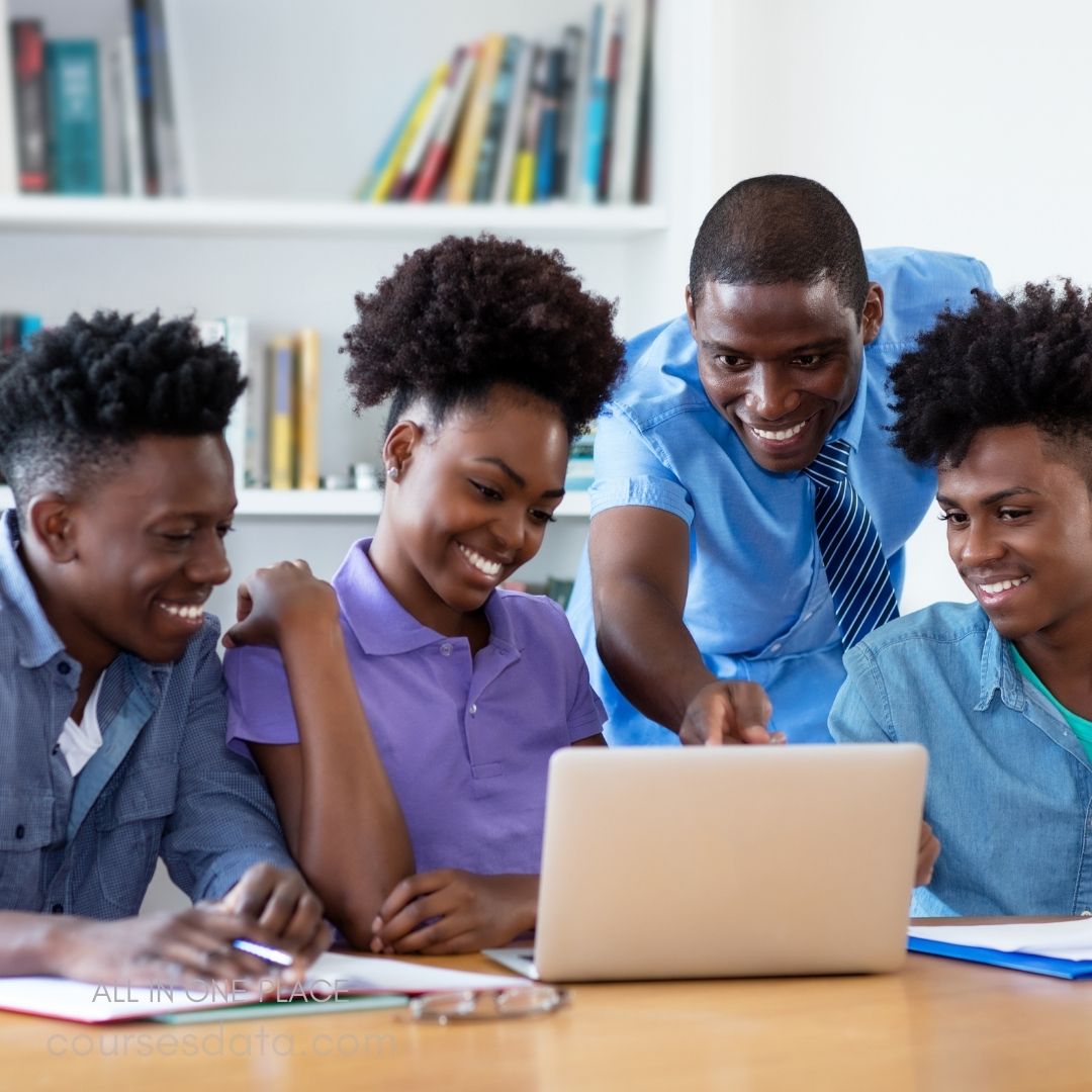Group studying around laptop. Smiling students engaged together. Teacher explaining concepts enthusiastically. Bookshelves filled with books behind. Colorful stationery on the table.