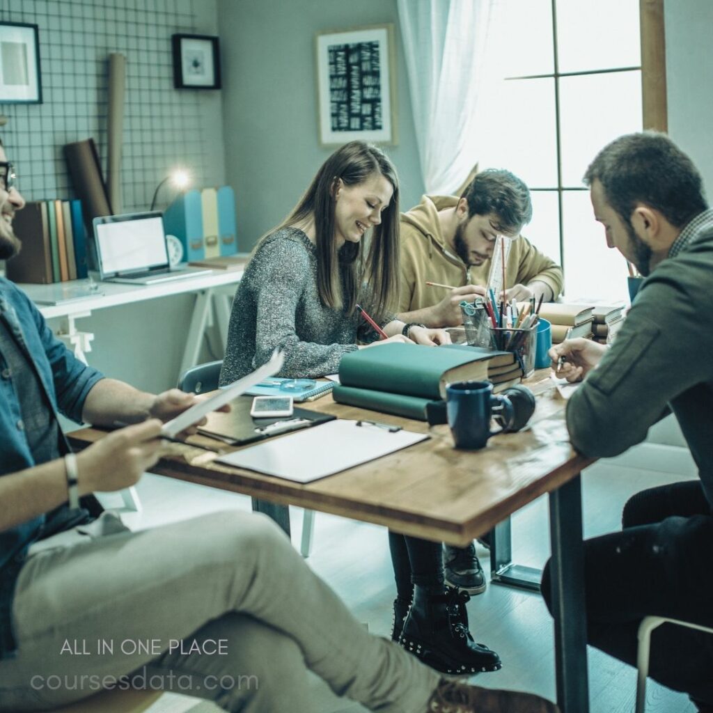 Group collaboration at wooden table. Four people engaged in discussion. Colorful stationery scattered on table. Laptop and books in background. Bright, cozy workspace atmosphere.