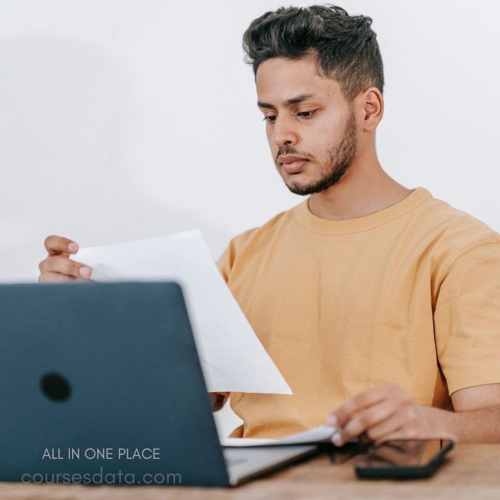 Man reading papers at desk. Laptop and smartphone present. Neutral background, focused expression. Casual orange t-shirt attire. Wooden surface, organized workspace.