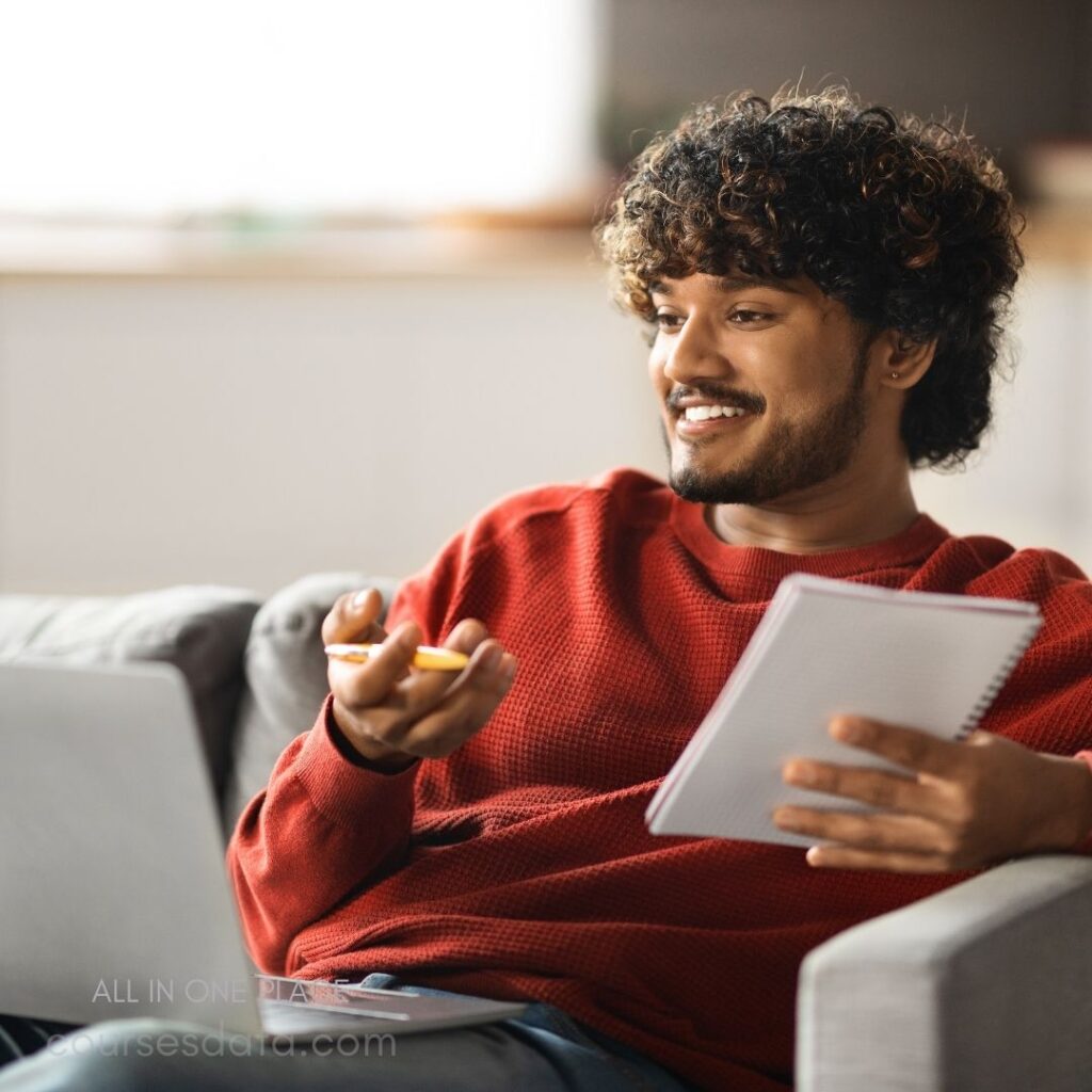 Smiling man with curly hair. Wearing a red sweater. Sitting on a couch. Holding a notebook and pen. Laptop open in front.