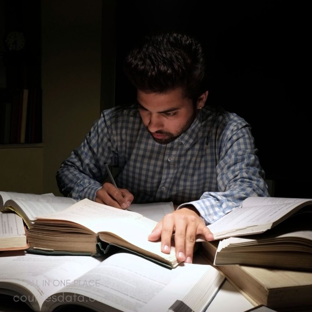 Student studying with books. Writing notes in dim light. Surrounded by open textbooks. Focused expression, deep concentration. Casual checkered shirt attire.