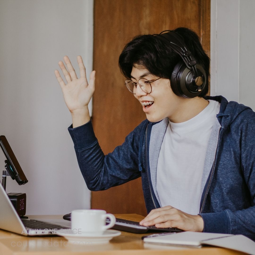 Young man waving happily. Wearing headphones and glasses. Sitting at a desk. Laptop and coffee cup nearby. Casual clothing, smiling expression.