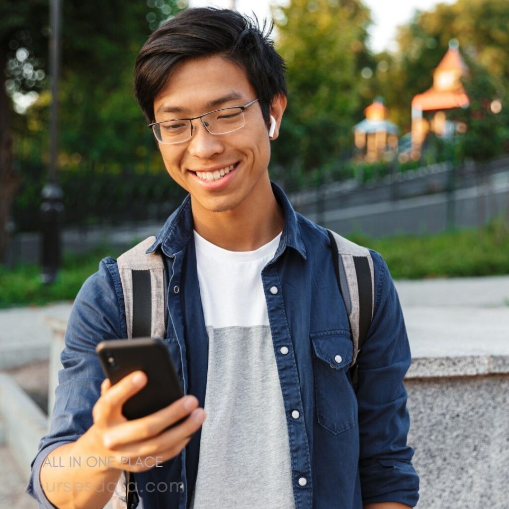 Smiling young man holding phone. Wearing glasses and earbuds. Casual outfit with backpack. Outdoor park setting. Sunny, lush background.