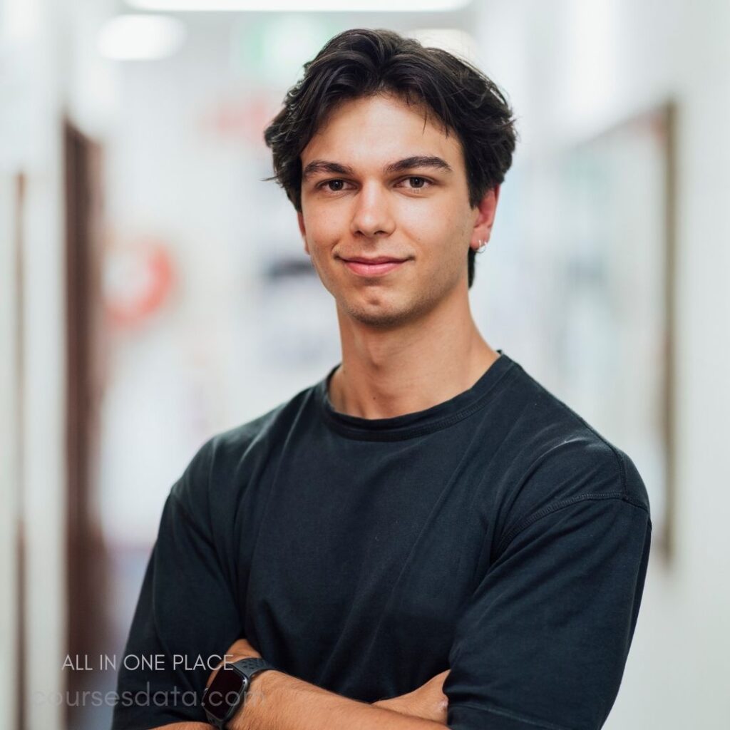 Young man with dark hair. Casual black T-shirt. Confident smile, arms crossed. Bright hallway background. Casual, professional appearance.