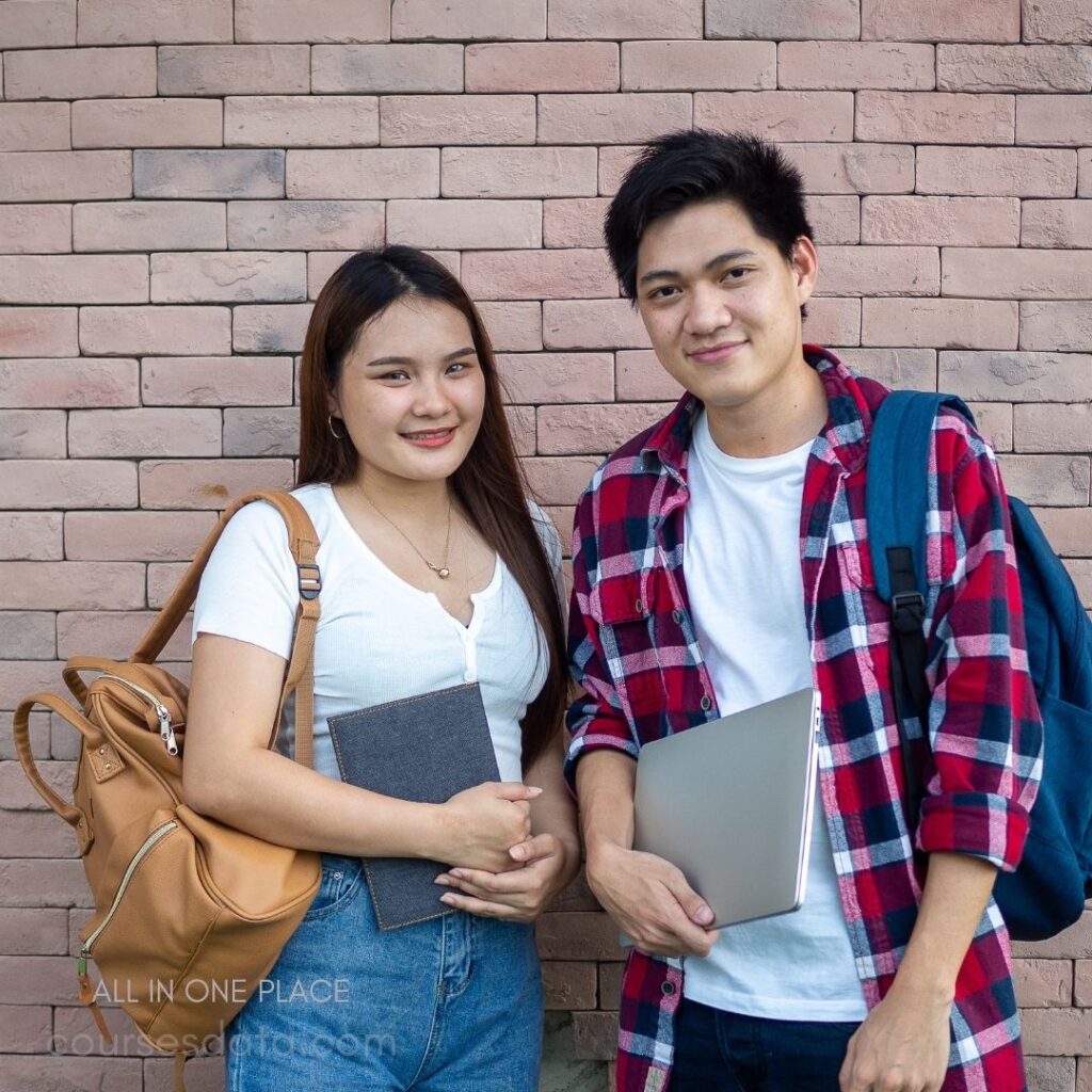 Two students smiling together. Tan backpack on woman. Male student holding a laptop. Brick wall background. Casual outfits, friendly atmosphere.