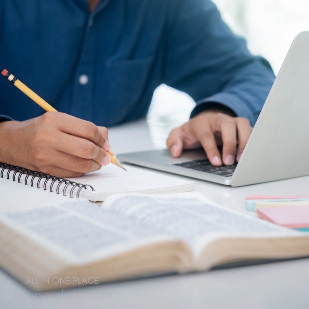 Person writing in notebook. Open laptop beside them. Textbook and sticky notes nearby. Casual blue shirt worn. Bright, natural light setting.