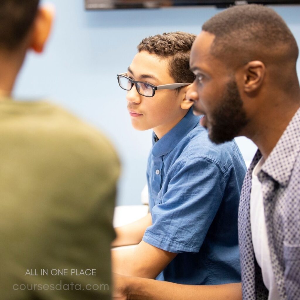 Young boy in blue shirt. Focused expression and glasses. Engaged in conversation. Adult man's profile visible. Classroom background.